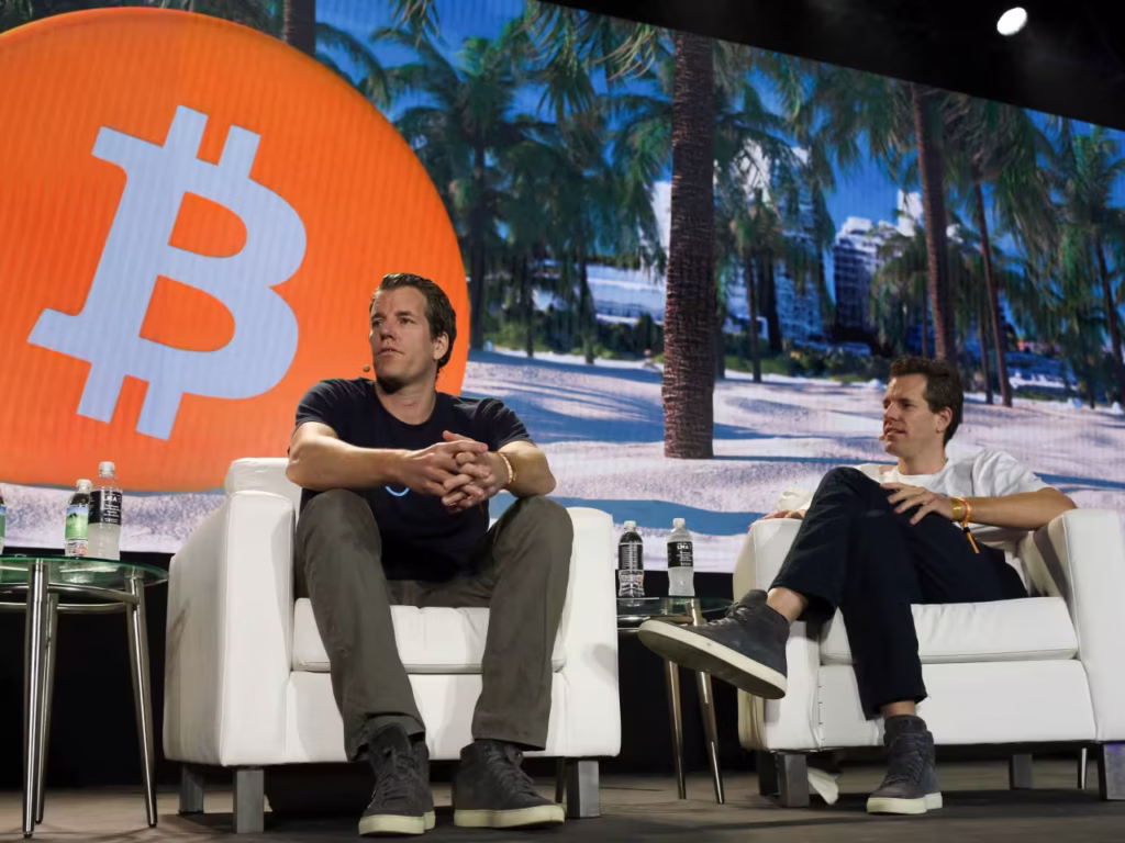 Cameron and Tyler Winklevoss, the co-founders of **Gemini**, are seated in white armchairs on a stage. Behind them is a large screen showing a tropical beach scene with a giant orange circle featuring the **Bitcoin** symbol.